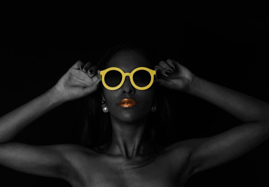 Artistic portrait of a woman wearing yellow sunglasses with gold lipstick accent.